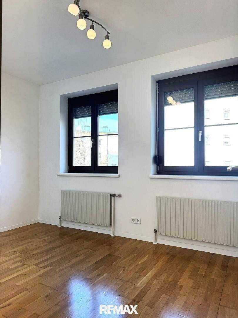 Heller Raum mit Parkettboden, zwei Fenstern mit dunklen Rahmen und Heizkörpern.