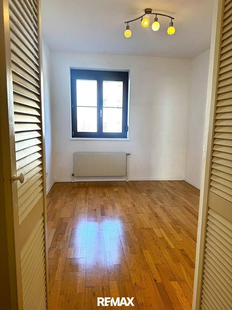 Leerer Raum mit Parkettboden und einem Fenster, gesehen durch offene Lamellentüren.