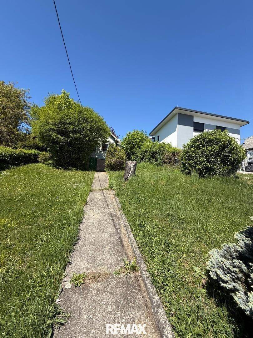 Betonweg führt durch eine grüne Wiese bergauf zu einem Haus, mit einem weiteren Gebäude im Hintergrund.