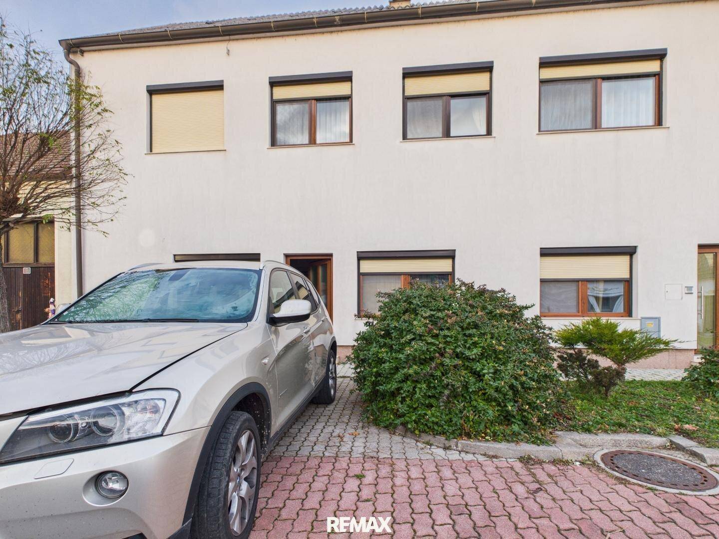 Vorderansicht des Hauses mit gepflastertem Parkplatz, grünen Büschen und Rollläden an den Fenstern.