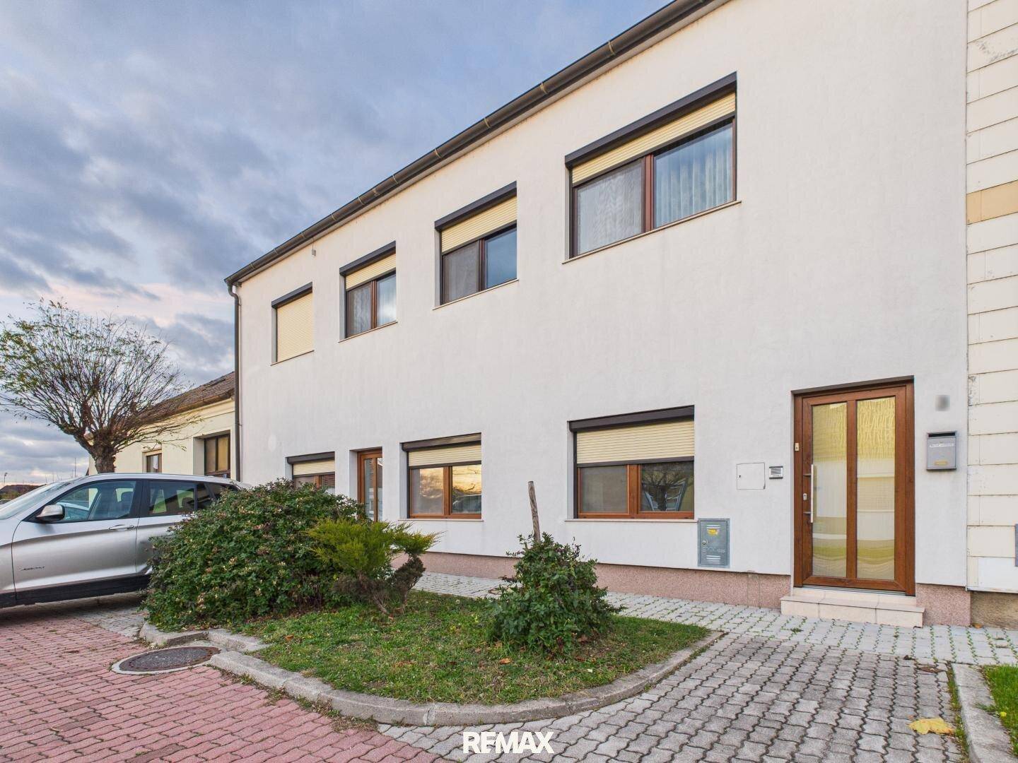 Weißes Mehrfamilienhaus mit braunen Fenstern und Rollläden, umgeben von einem kleinen Vorgarten.