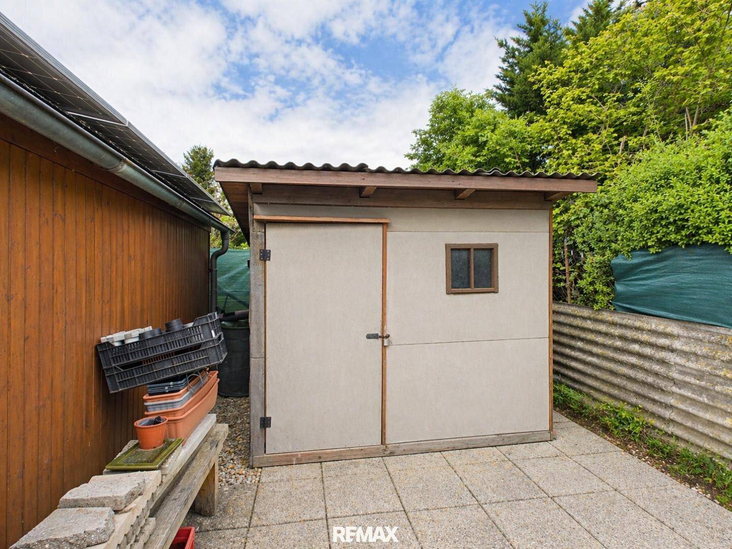 Kleiner Gartenschuppen mit Wellblechdach und gepflastertem Vorplatz, umgeben von üppigem Grün.