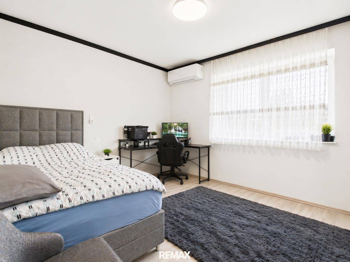 Schlafzimmer mit Doppelbett, Schreibtisch und Klimaanlage, hell durch großes Fenster.