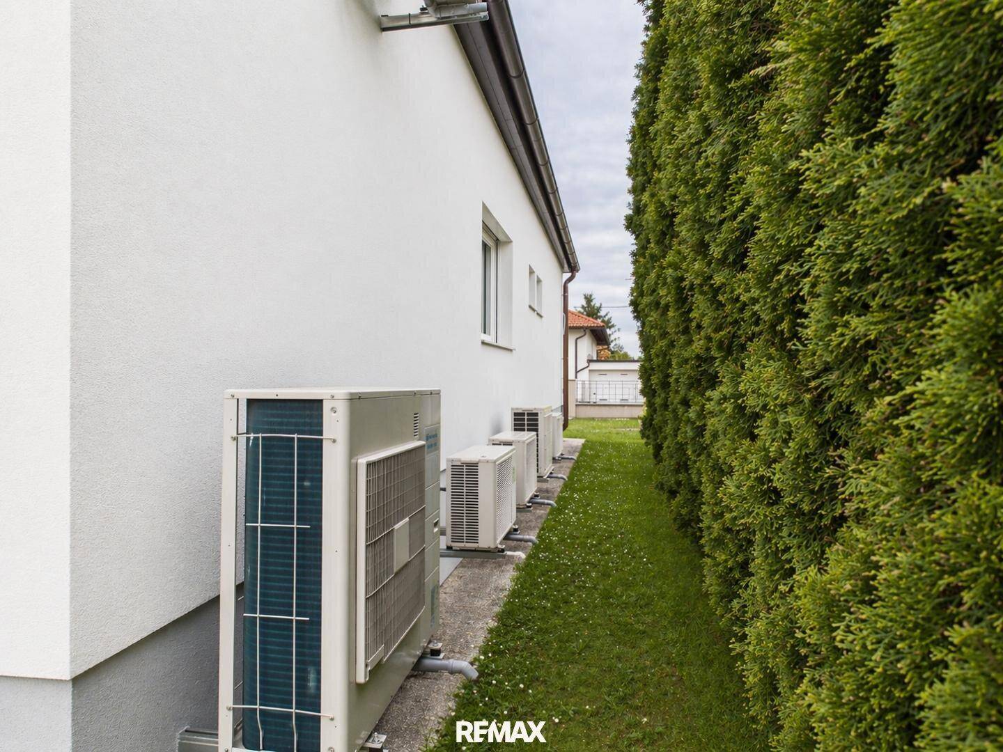 Reihe von Klimaanlagen-Außengeräten entlang der Hauswand neben einer hohen Hecke.