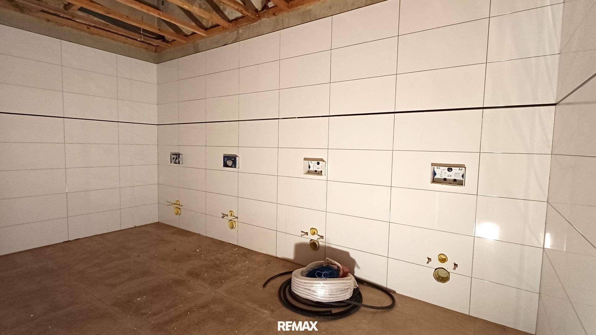 Badezimmer im Rohbau mit weißen Wandfliesen und vorbereiteten Anschlüssen für Sanitäranlagen.