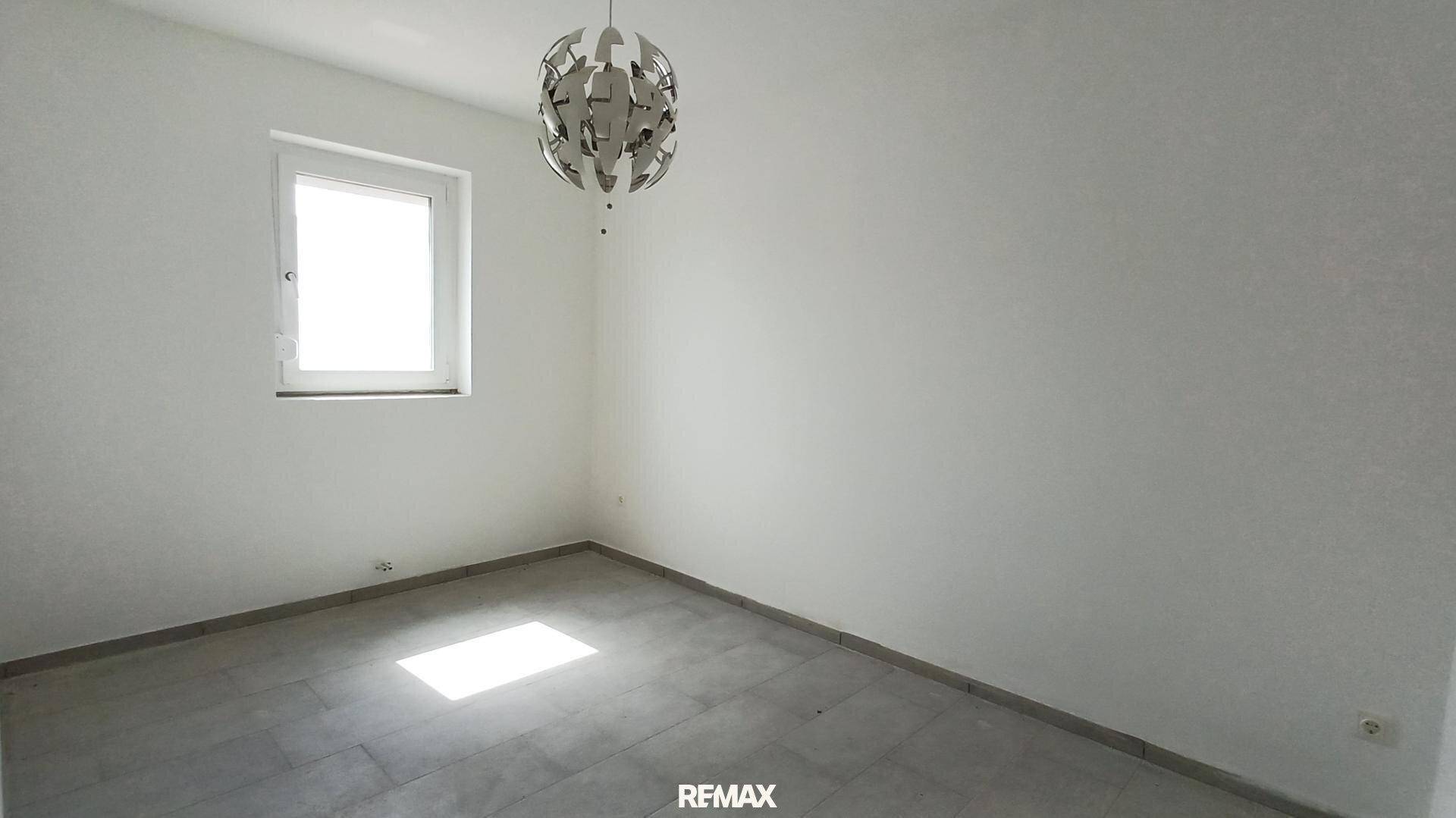 Helles, leeres Zimmer mit einem Fenster und einer modernen Deckenleuchte, bereit zur Einrichtung.