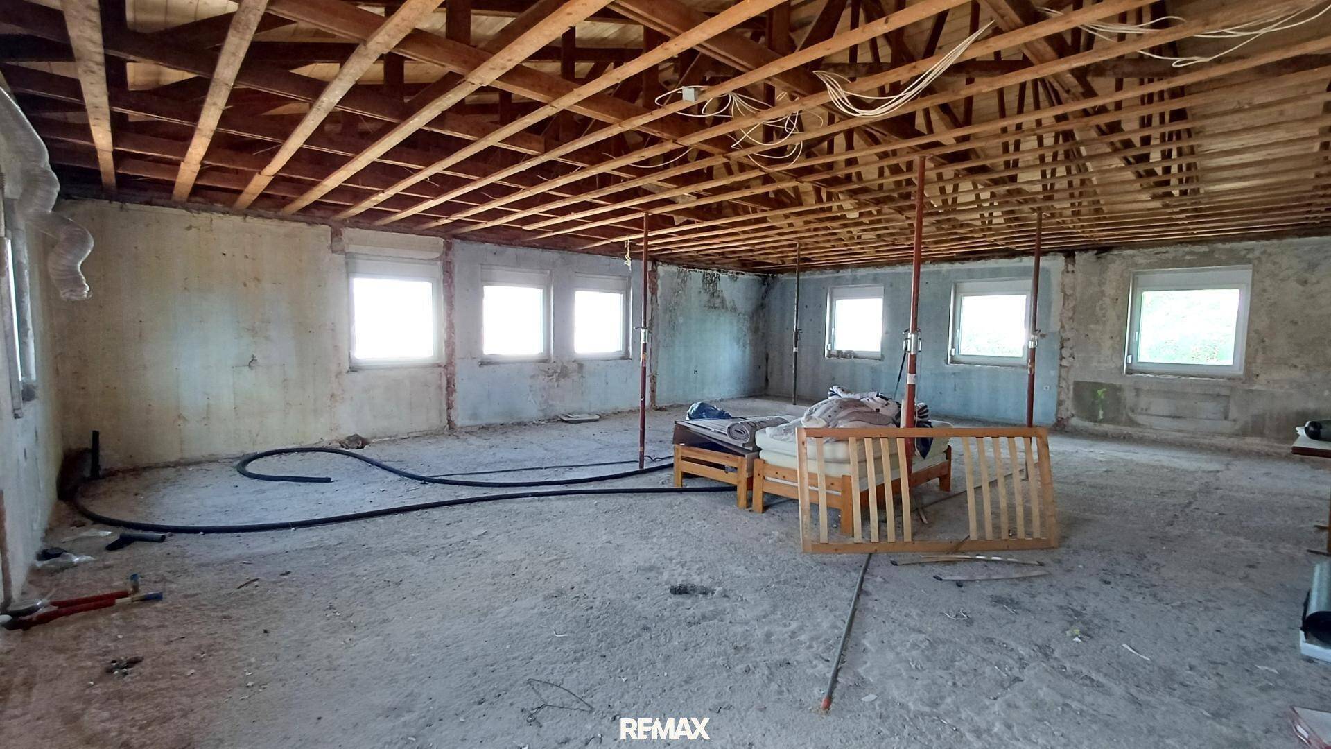 Großer Raum im Rohbau mit sichtbaren Holzbalken und mehreren Fenstern, viel Potenzial.