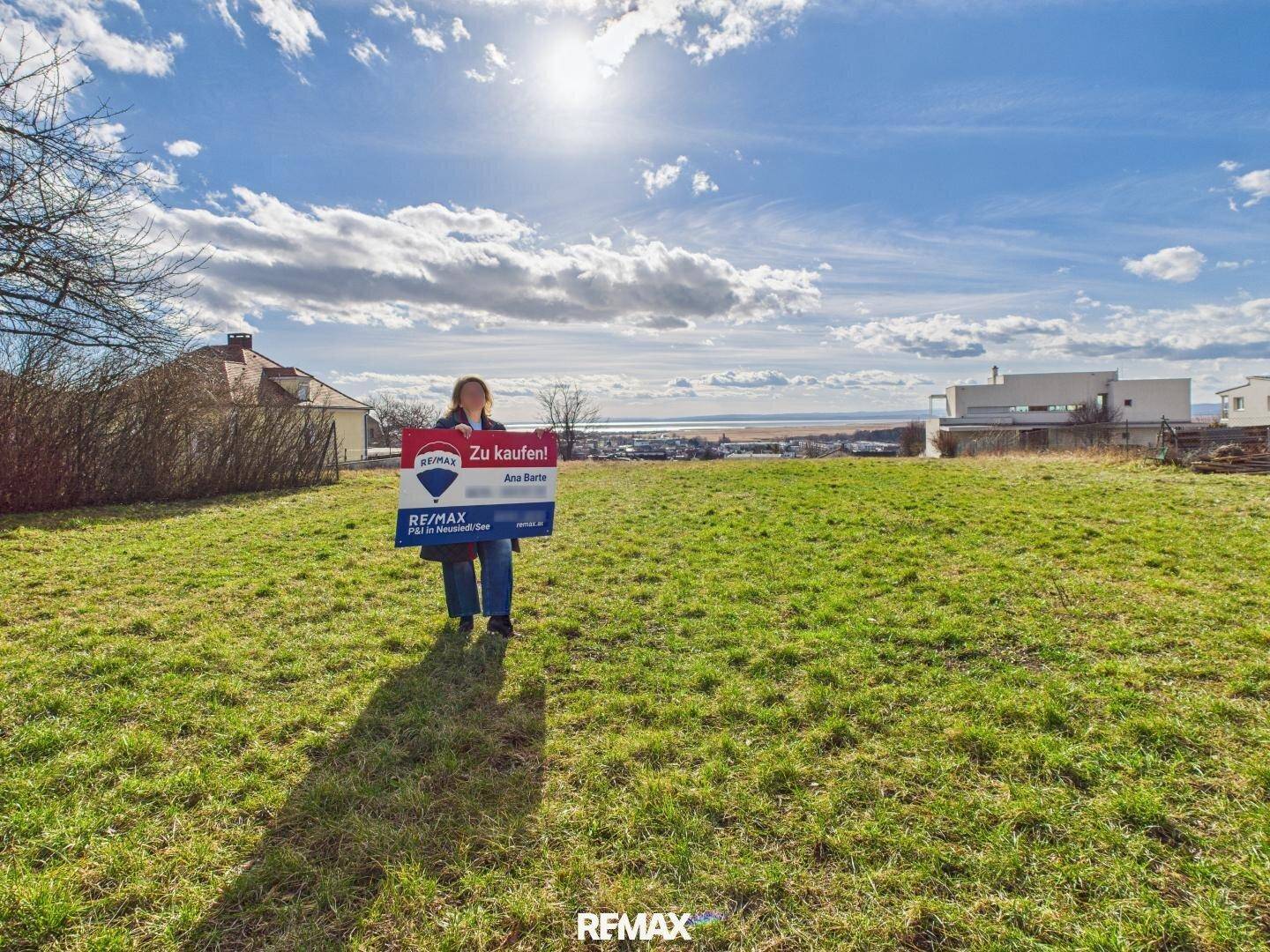 Sonnenverwöhntes Grundstück mit grüner Wiese, Verkaufsschild und Panoramablick auf den See.