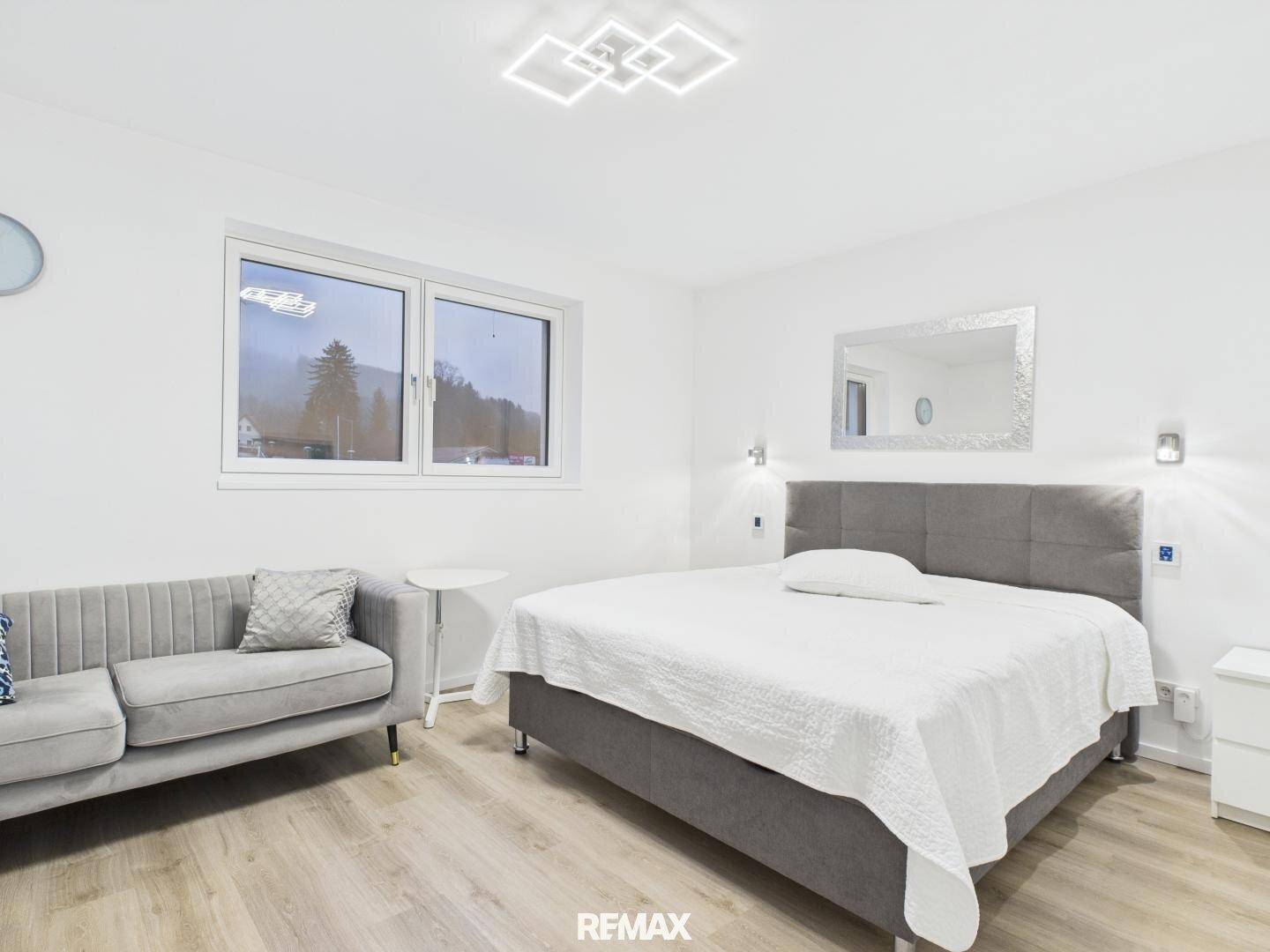 Helles Schlafzimmer mit modernem Bett, Sofa und Blick auf die Landschaft durch das Fenster.