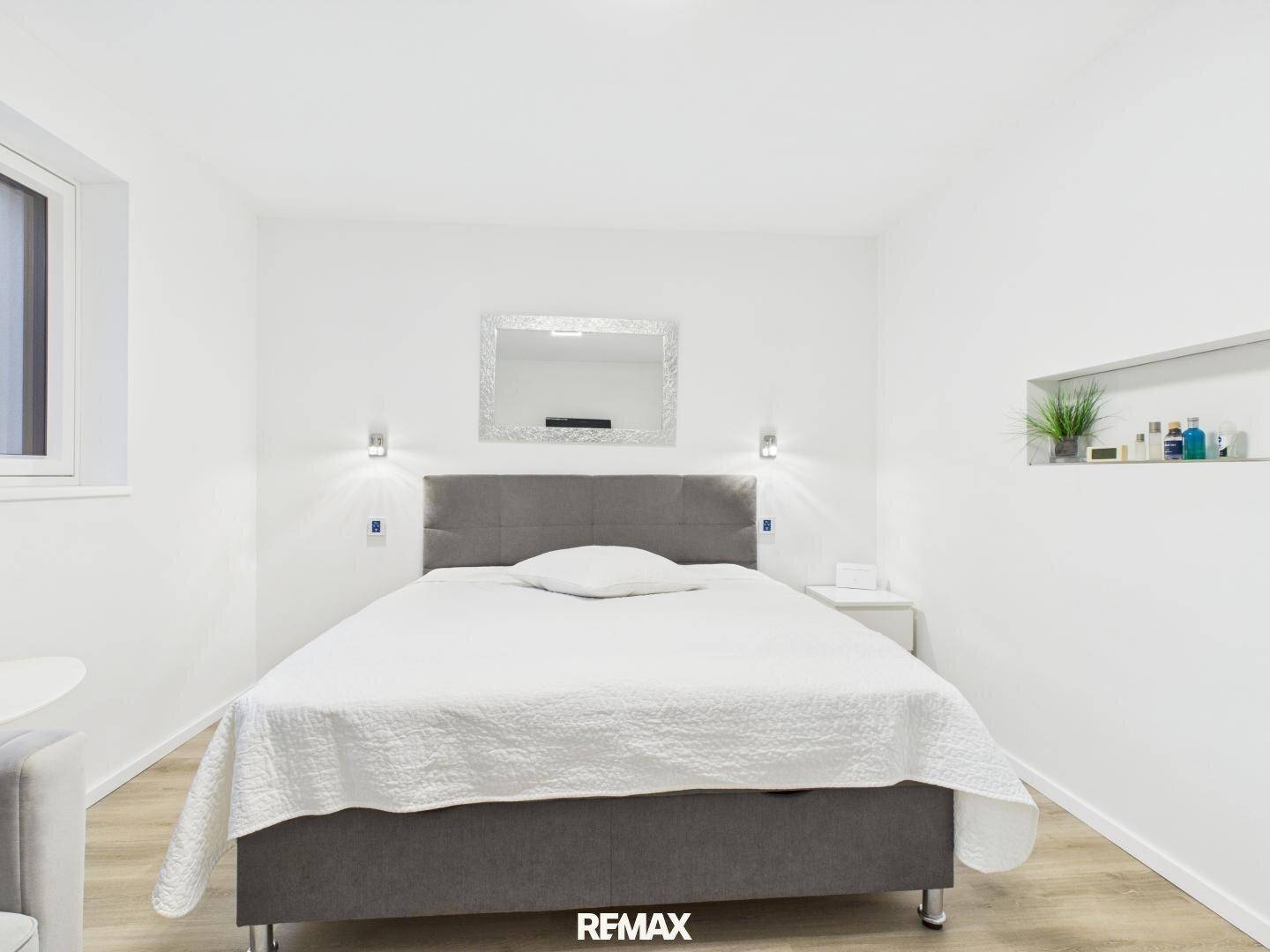 Modernes Schlafzimmer mit grauem Polsterbett, Wandspiegel und integrierten Ablagen.