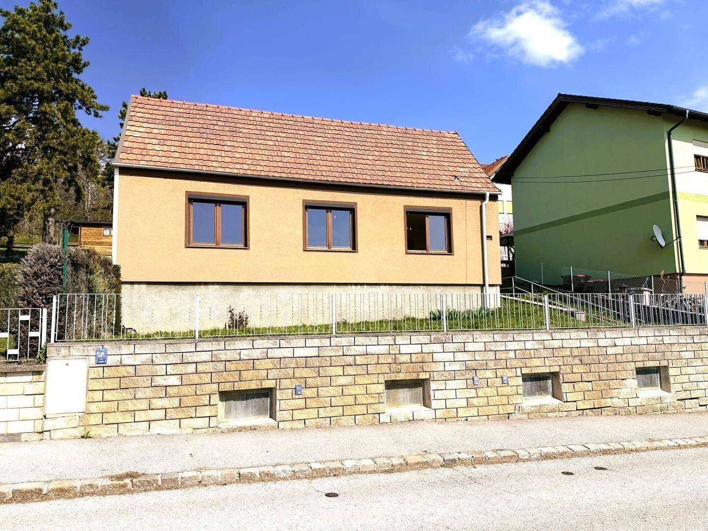 Vorderansicht eines Einfamilienhauses mit beiger Fassade und braunen Fenstern, umgeben von einer Steinmauer.