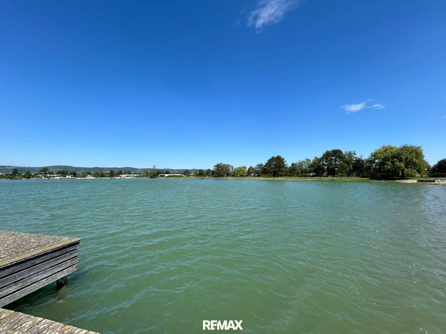 Weitläufiger Blick über einen ruhigen See mit Holzsteg und bewaldeten Ufern.