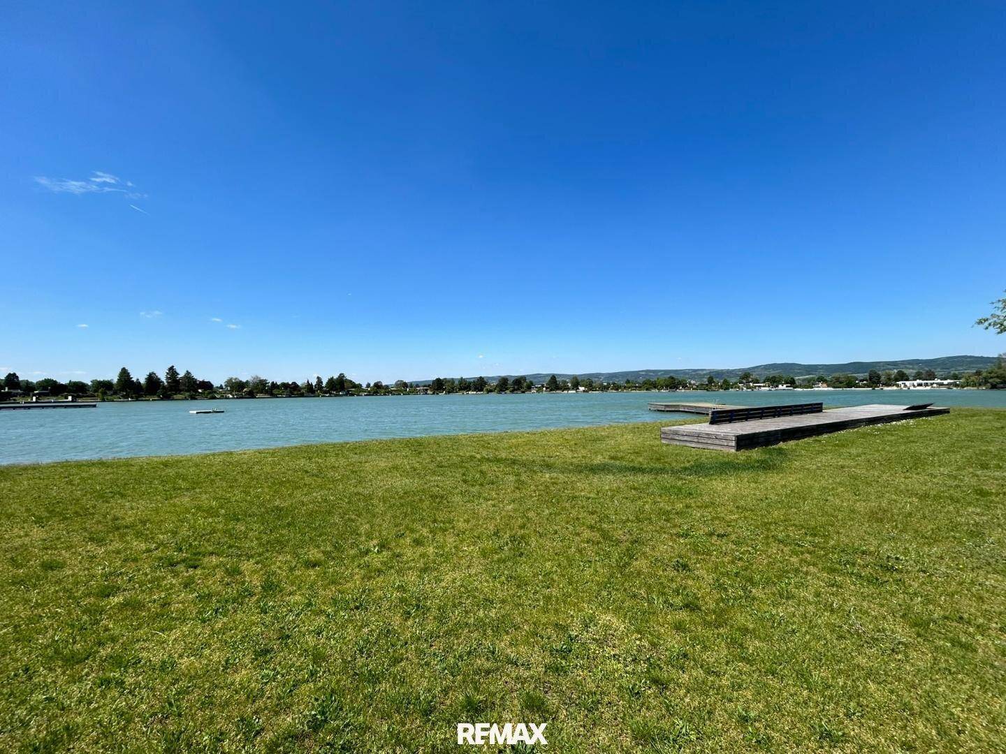 Panoramablick auf den See mit grünen Wiesen im Vordergrund und klarem Himmel.