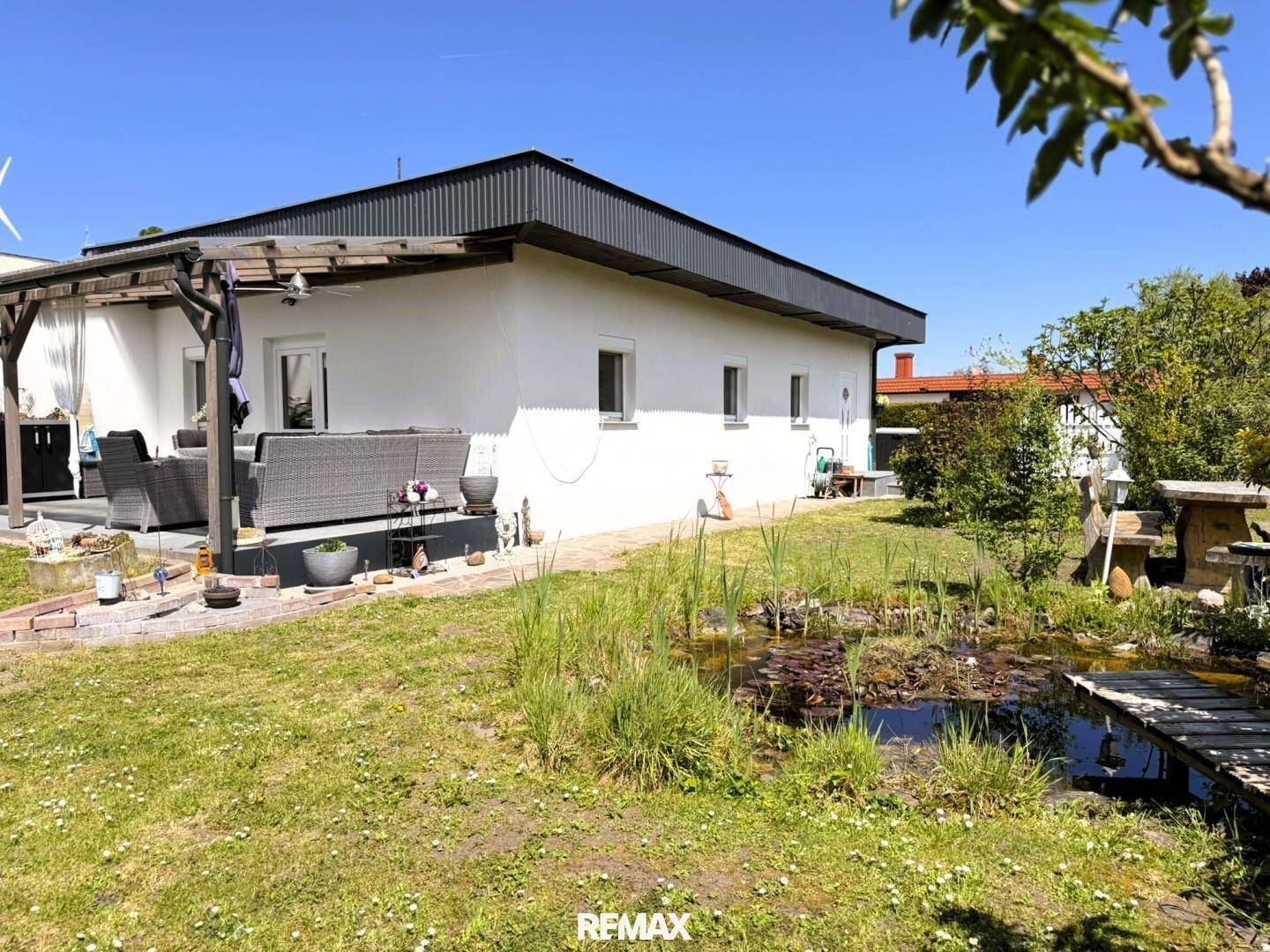 Rückseite des Hauses mit Terrasse, Teich und weitläufigem Garten unter blauem Himmel.