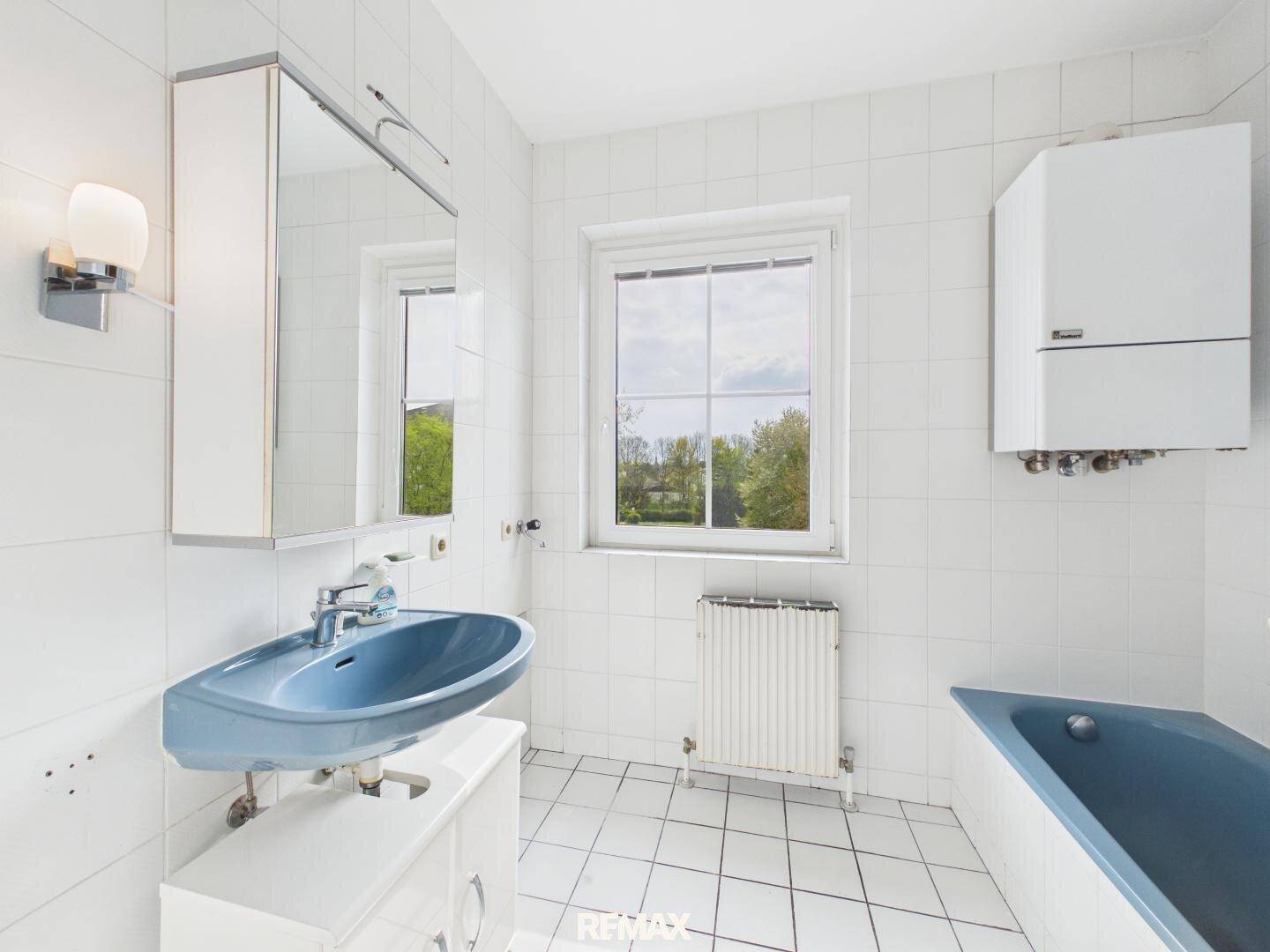 Badezimmer mit blauer Badewanne, Waschbecken und Fenster, gefliest in Weiß.