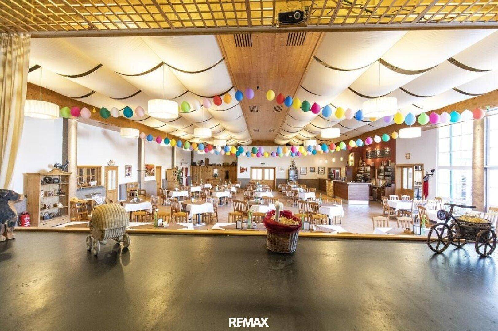 Weitläufiger Festsaal mit Bühne, vielen Tischen und bunter Ballondekoration, ideal für große Feiern.