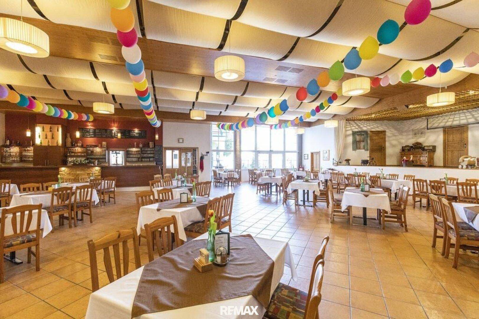 Großer, festlich dekorierter Saal mit vielen Sitzgelegenheiten und bunten Ballons an der Decke.