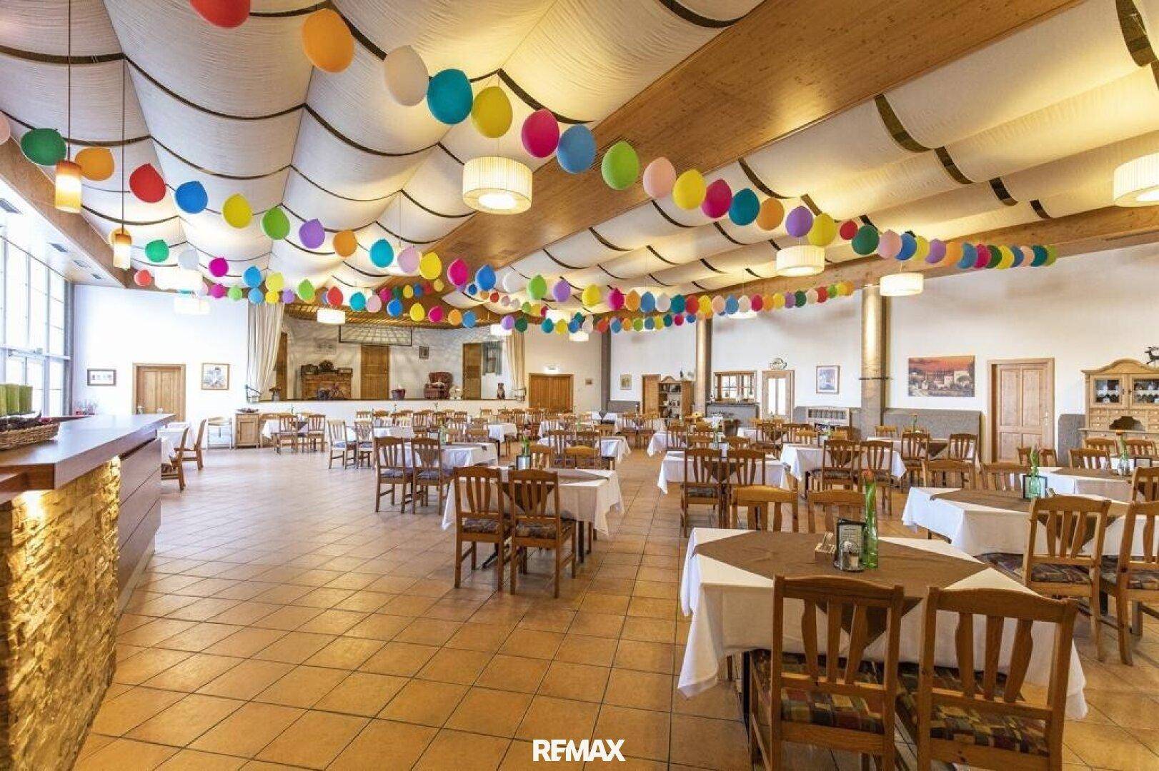 Großer Festsaal mit vielen Tischen, Stühlen und bunter Ballondekoration, ideal für Feierlichkeiten.