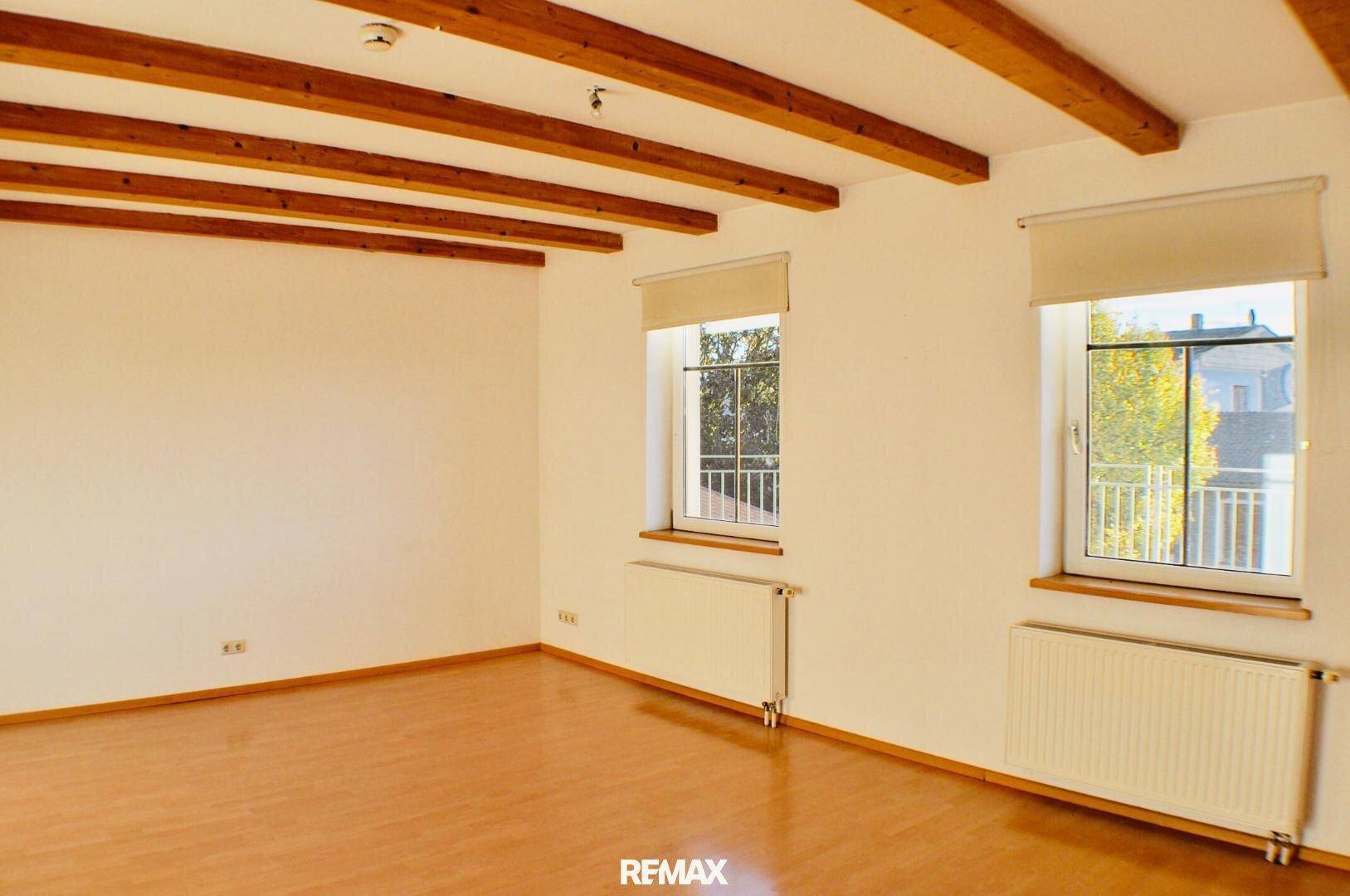 Heller Wohnraum mit sichtbaren Holzbalken, Laminatboden und zwei Fenstern, gemütliche Atmosphäre.