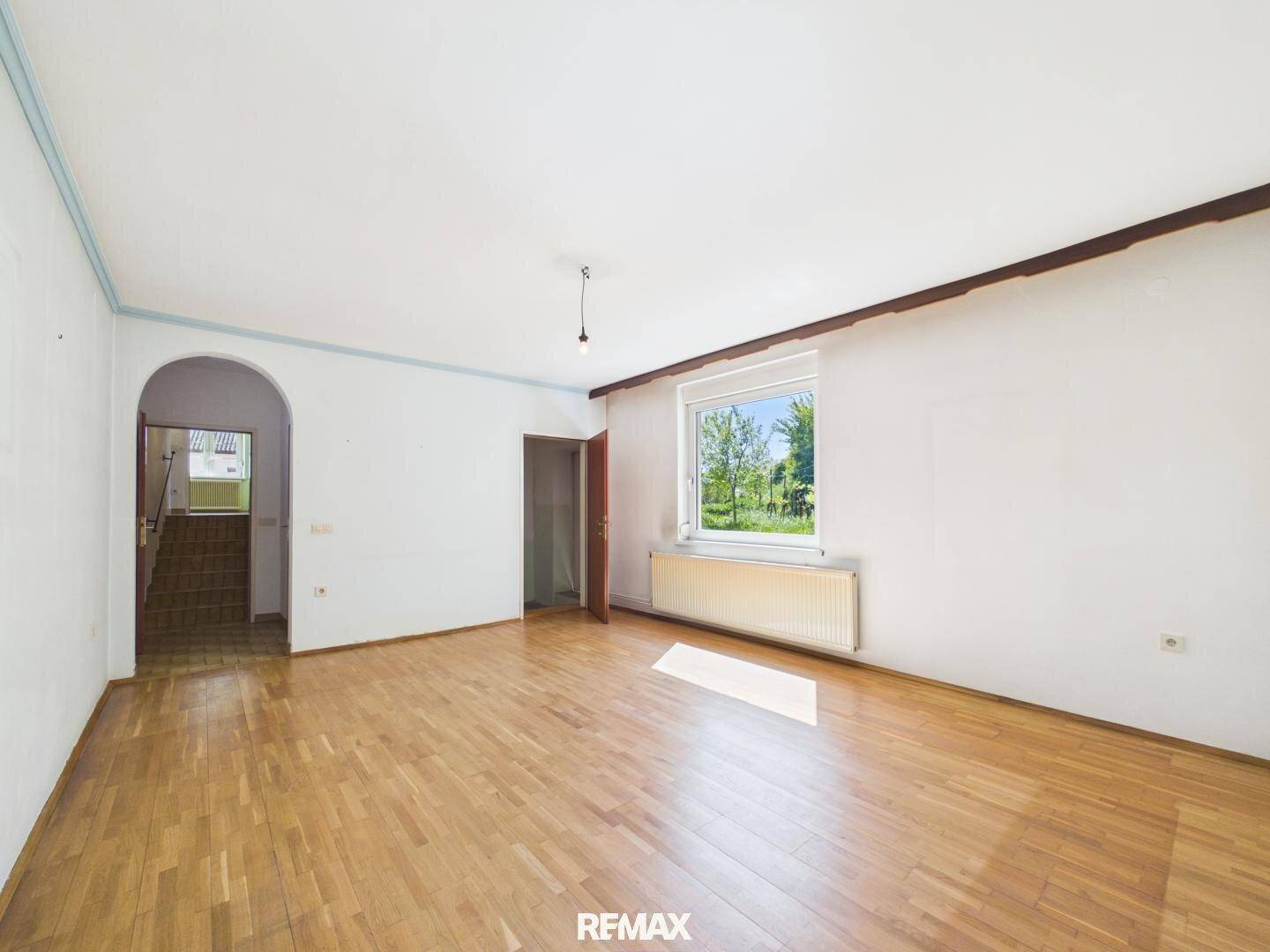 Helles Wohnzimmer mit Parkettboden und einem Fenster, das einen Blick ins Grüne bietet.