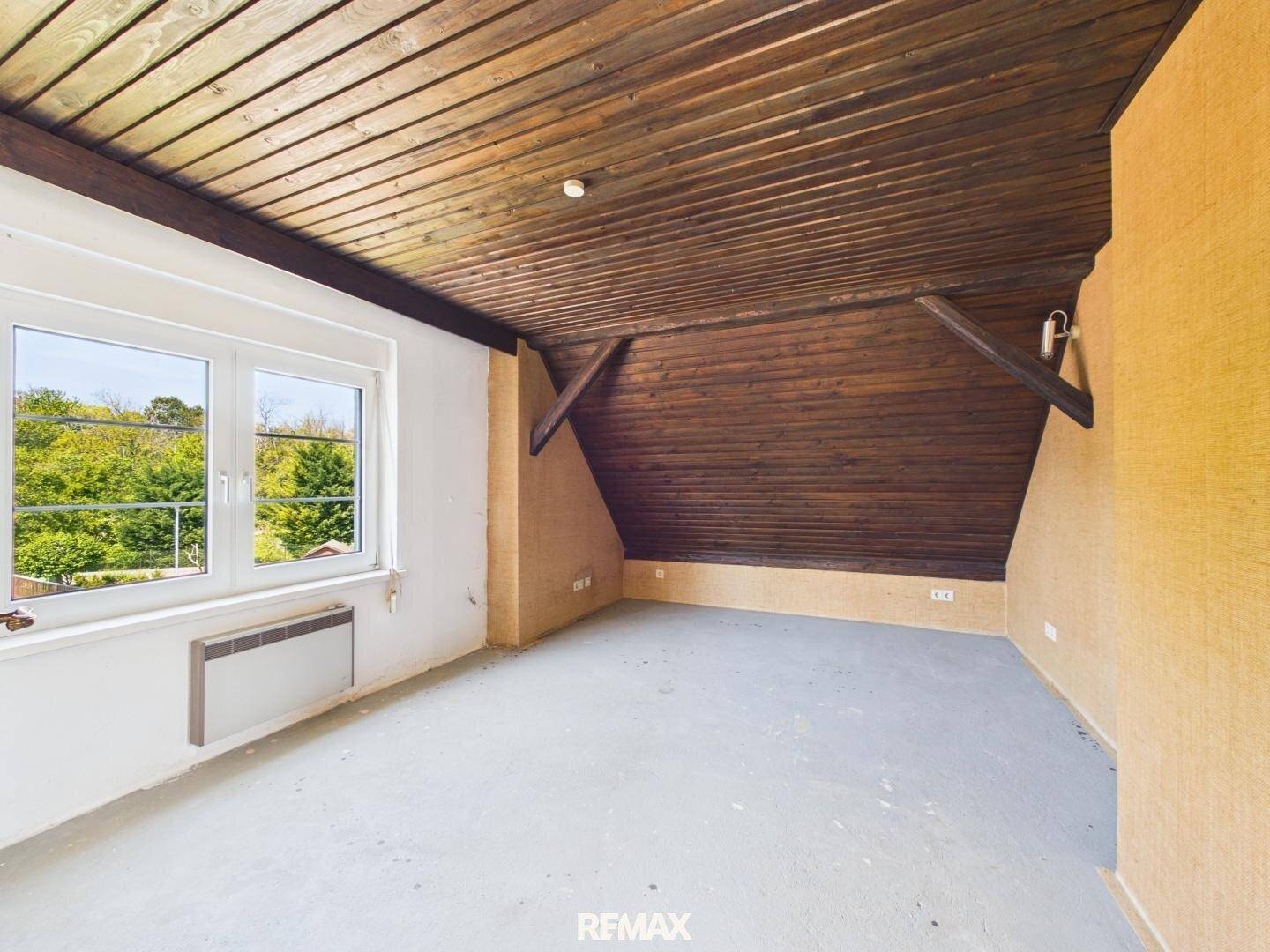 Dachzimmer mit Holzverkleidung an Decke und Wänden, sowie einem Fenster mit Blick ins Grüne.