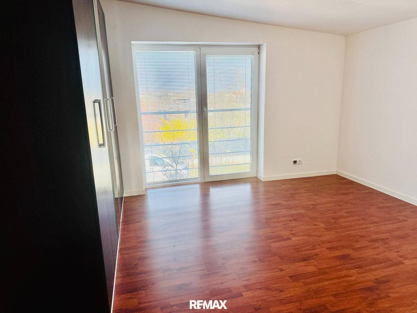 Helles Schlafzimmer mit dunklem Laminatboden und großen Fenstern mit Balkonzugang.