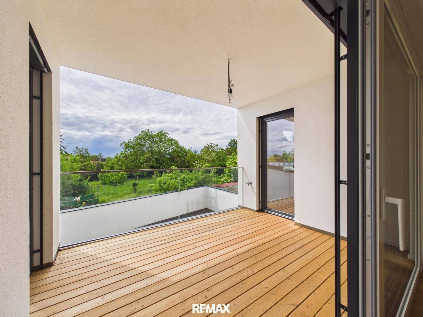Geräumiger Balkon mit Holzdielenboden und Glasgeländer, umgeben von viel Grün.