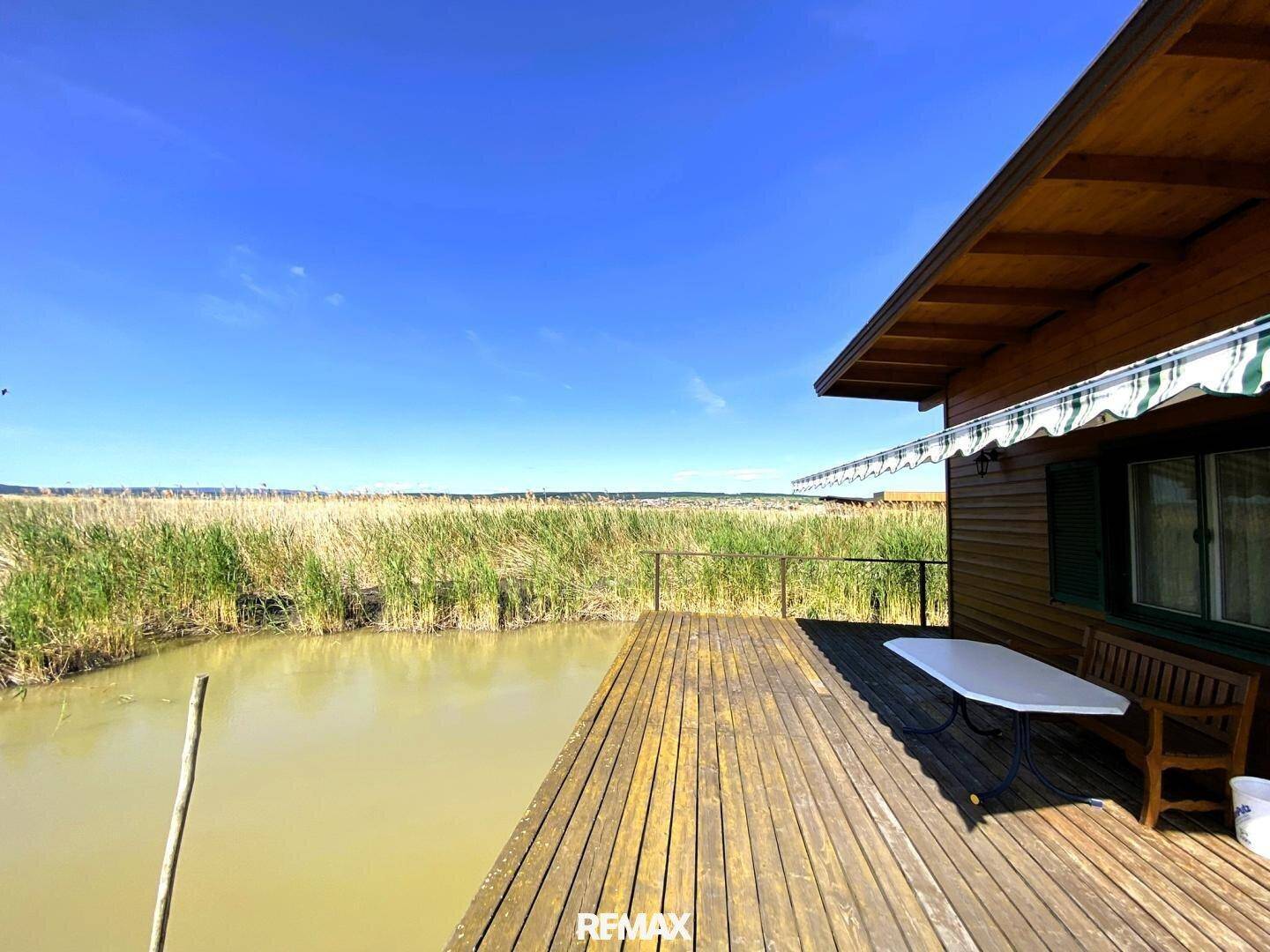 Geräumige Holzterrasse mit Tisch und Bank, bietet einen weiten Blick über das Wasser und Schilf.