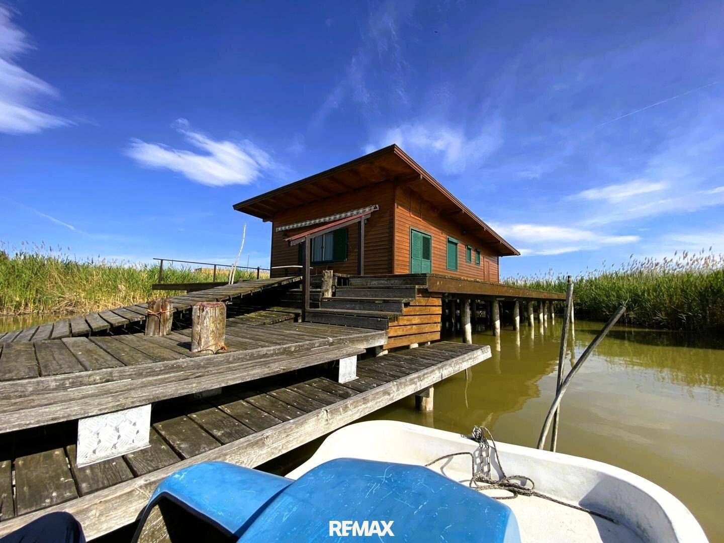Holzhaus auf Stelzen über dem Wasser, von einem Boot aus gesehen, mit einer weitläufigen Holzterrasse.
