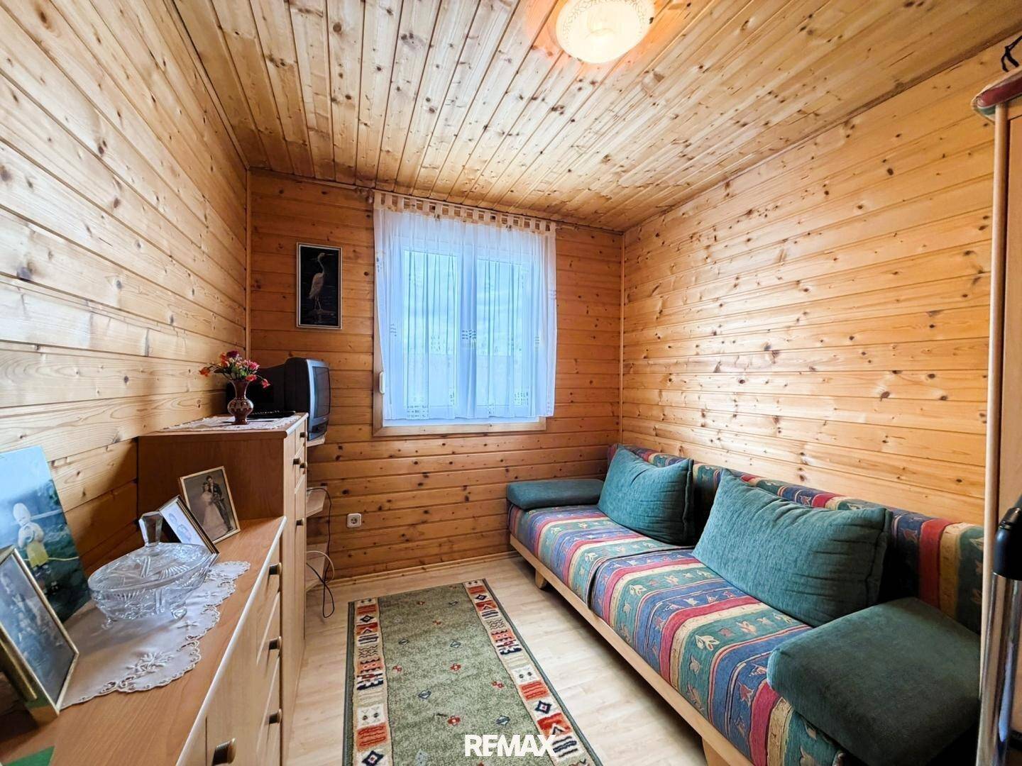 Kleines Schlafzimmer mit einem Sofa, Holzwänden und -decke, sowie einem Fenster mit Vorhängen.