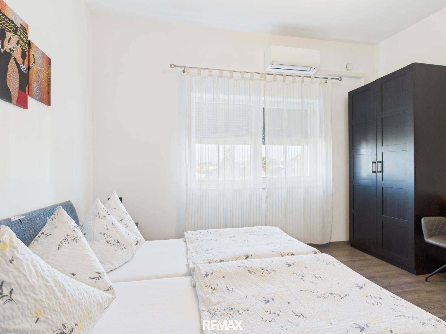 Helles Schlafzimmer mit zwei Einzelbetten, einem großen Kleiderschrank und einem Fenster mit Vorhängen.