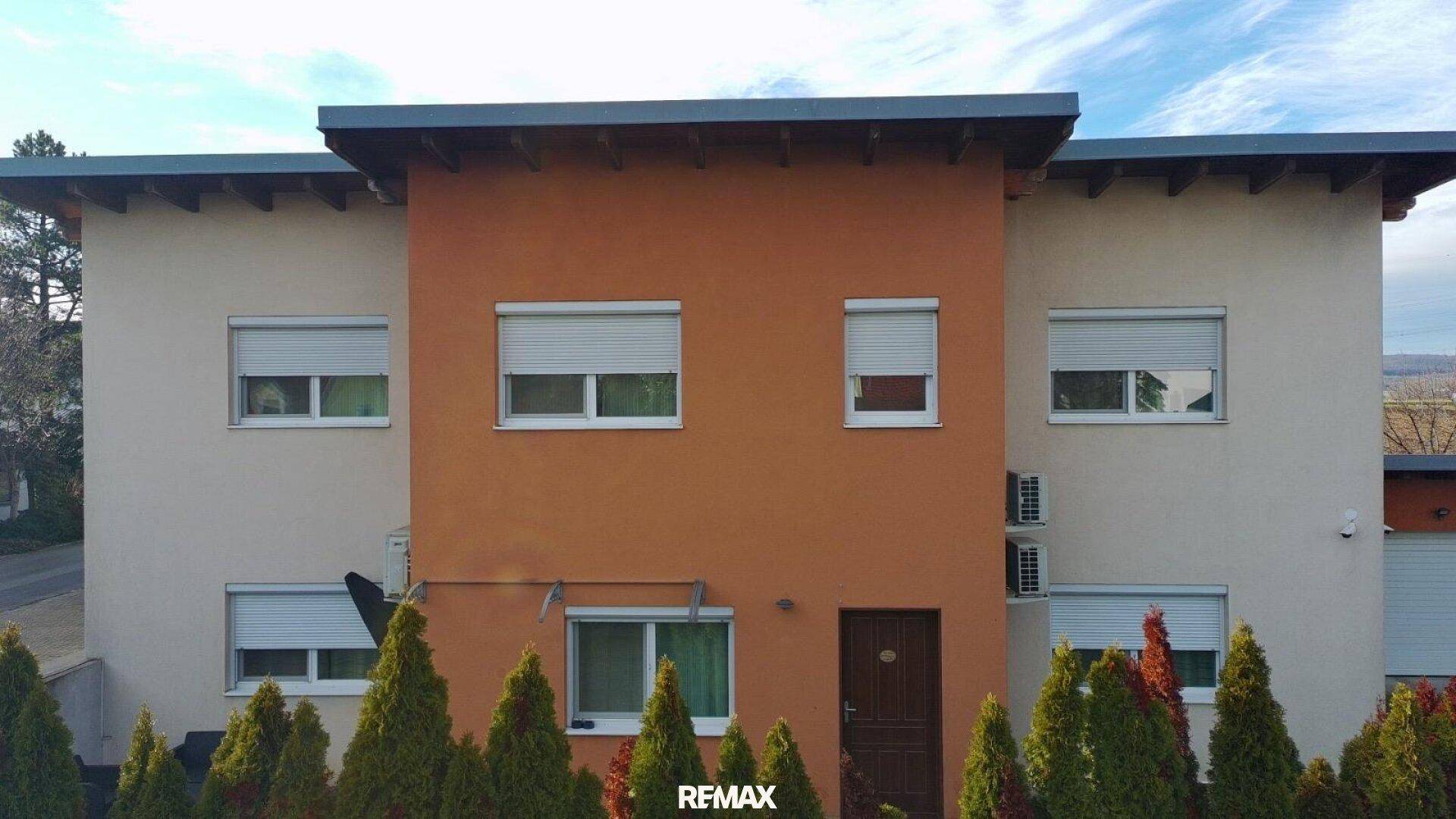 Vorderansicht des modernen Hauses mit beiger und oranger Fassade sowie mehreren Fenstern mit Rollläden.