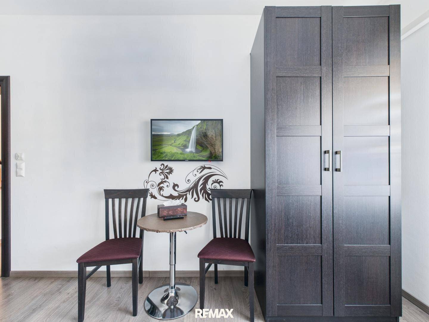 Kleiner Sitzbereich mit Tisch und Stühlen, einem Fernseher und einem dunklen Kleiderschrank.