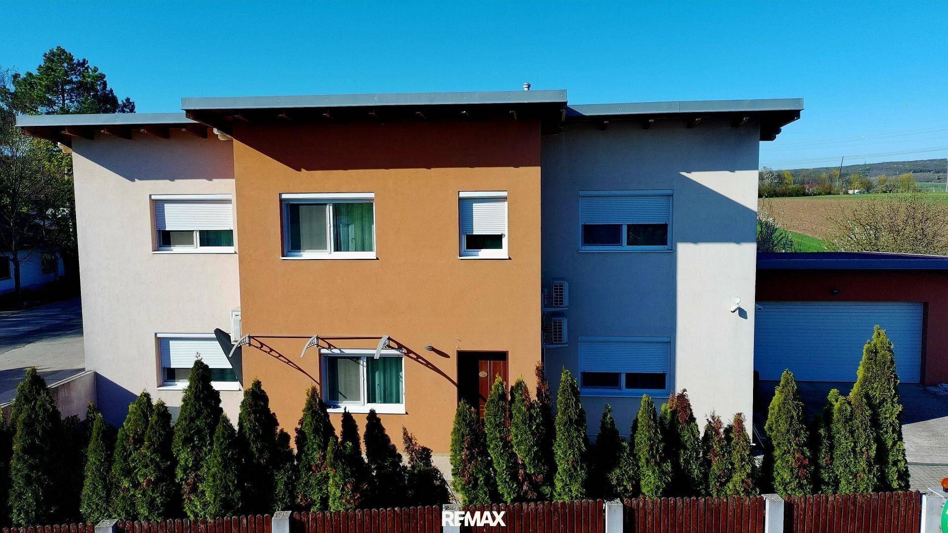 Modernes zweistöckiges Haus mit flachem Dach, weißer und orangefarbener Fassade sowie Rollläden an den Fenstern.