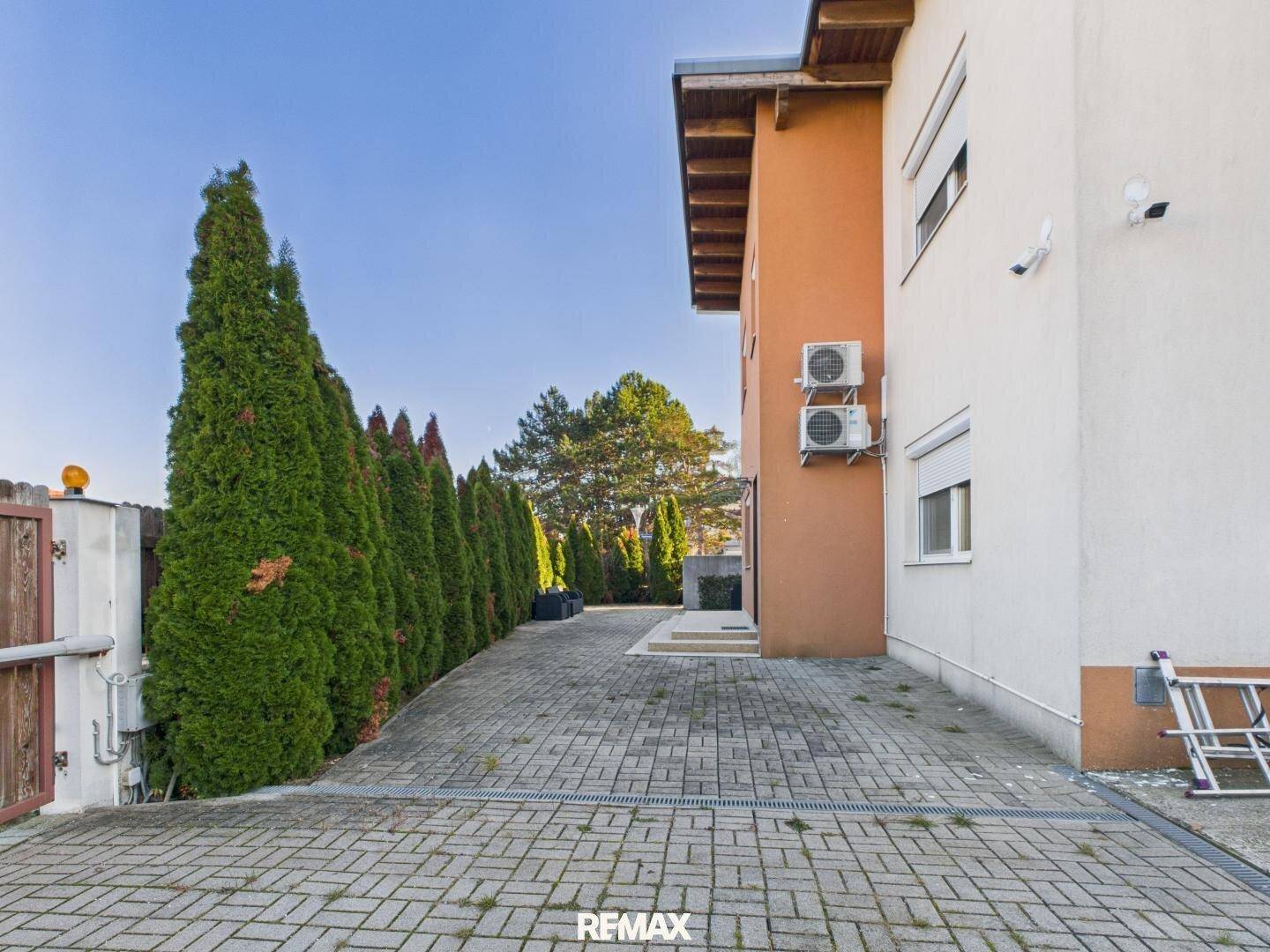 Seitenansicht des Hauses mit langer gepflasterter Einfahrt und einer hohen Hecke als Begrenzung.