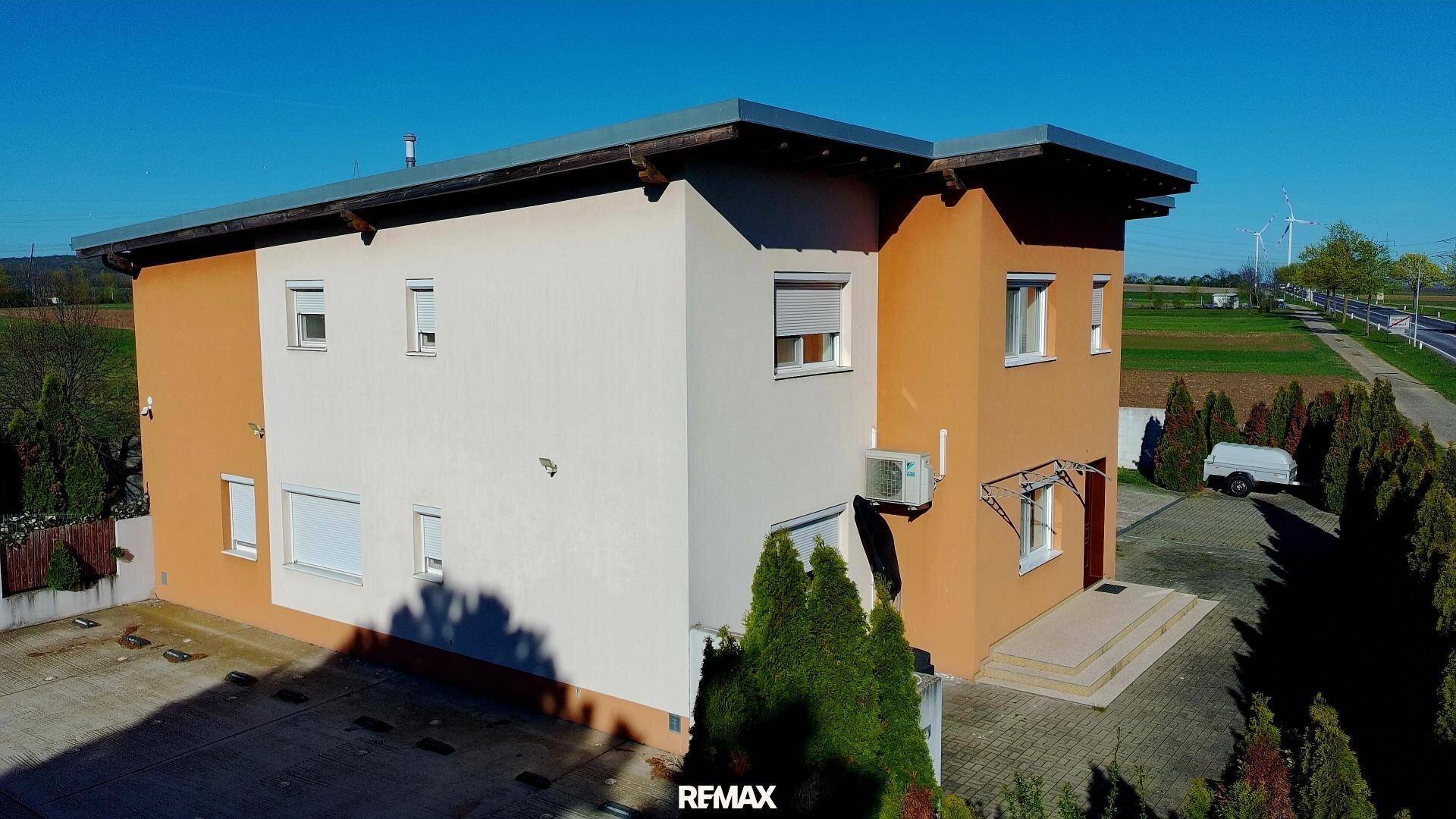 Seitenansicht eines modernen Hauses mit großem Vorplatz, umgeben von Bäumen und Feldern in der Ferne.