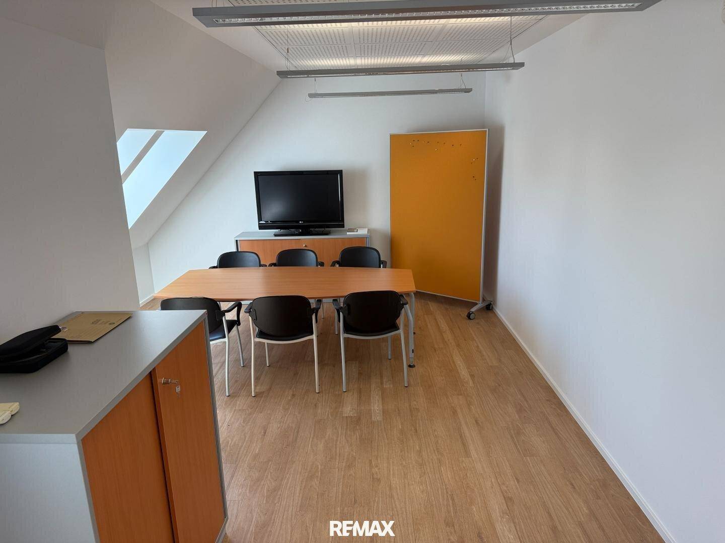 Heller Büroraum mit Dachschräge, Holzboden und einem Besprechungstisch unter dem Dachfenster.