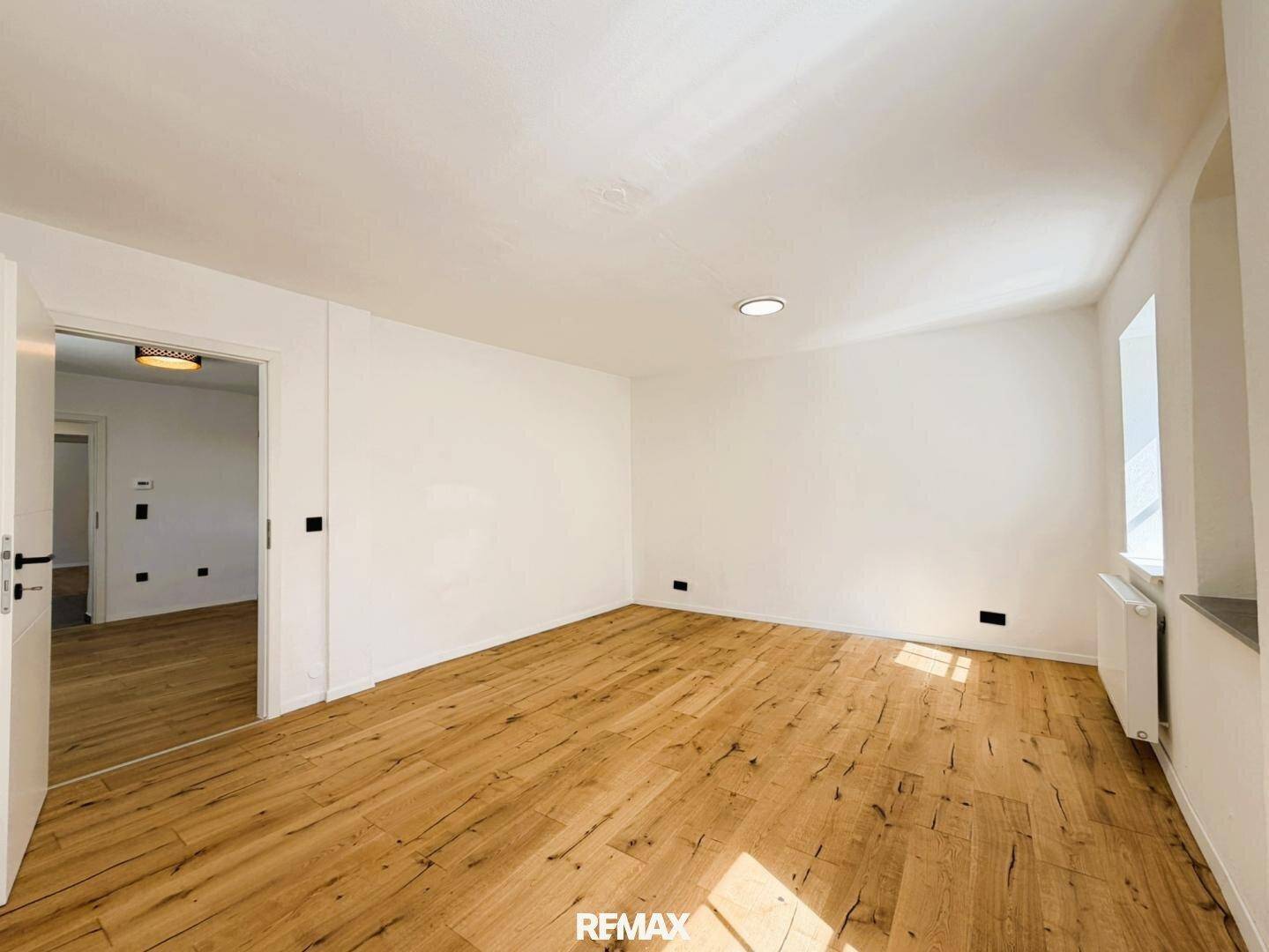 Lichtdurchfluteter Raum mit Holzboden und weißen Wänden, bietet Zugang zu einem weiteren Zimmer.