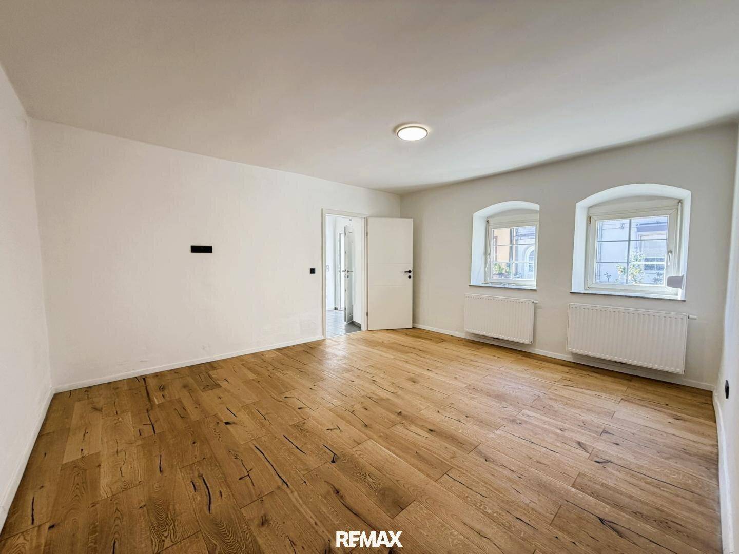 Geräumiger Wohnraum mit Holzboden und weißen Wänden, bietet viel natürliches Licht.