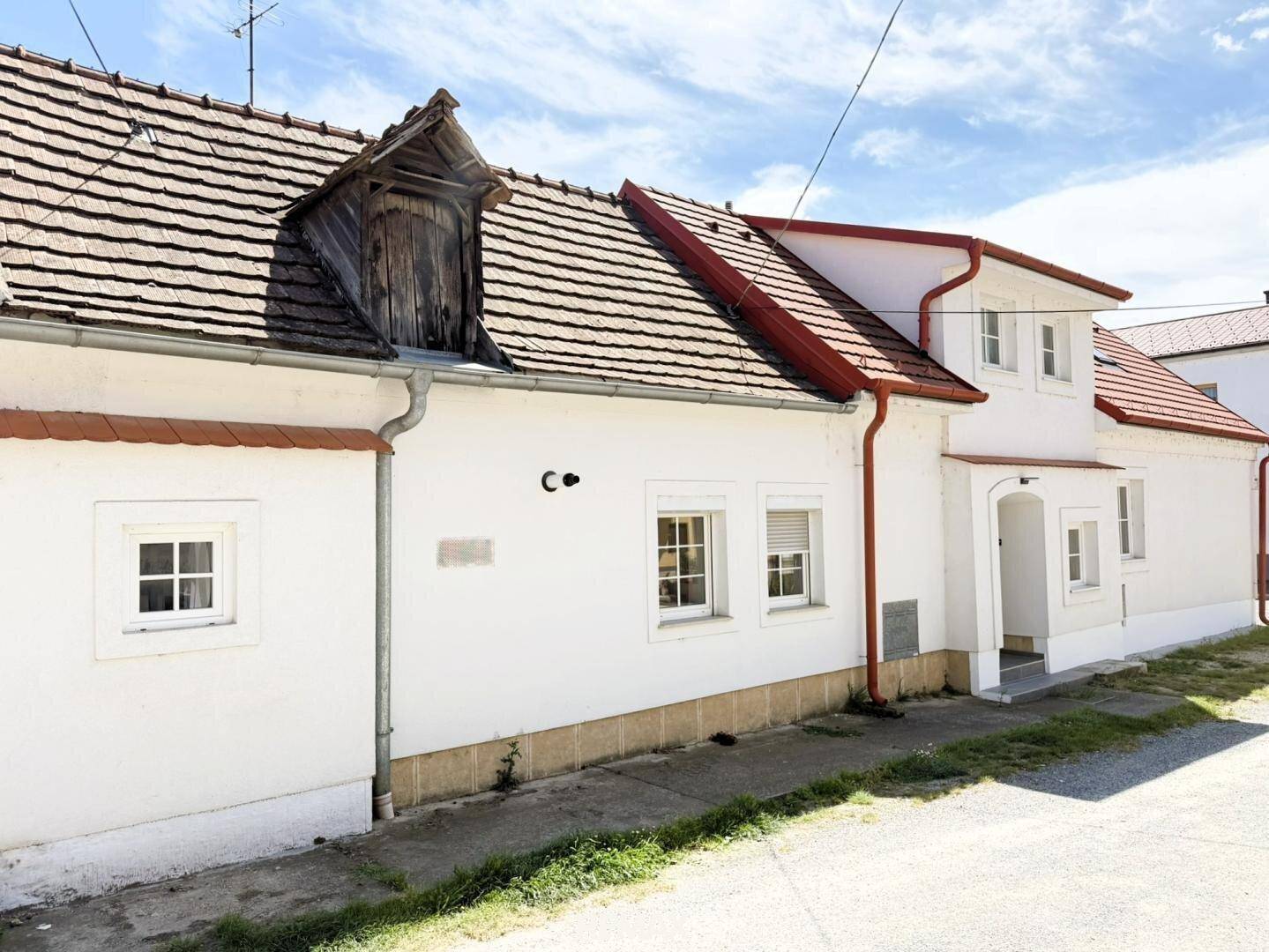 Längere Ansicht des weißen Hauses mit rotem Ziegeldach und einem alten Dachgaubenfenster.