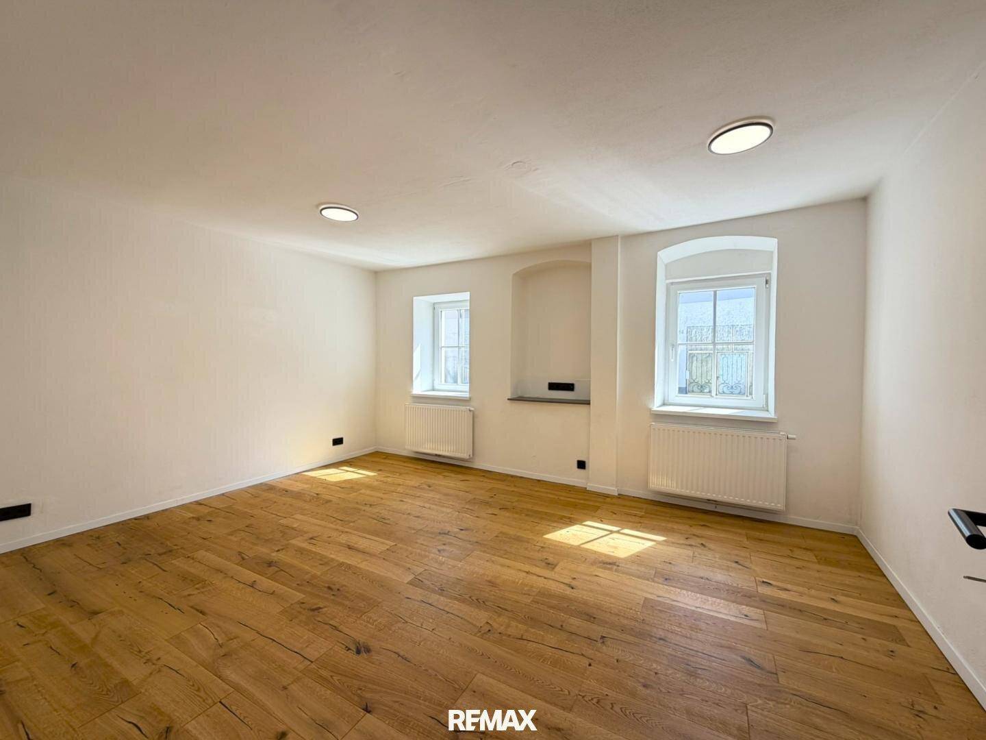 Heller Wohnraum mit Holzboden, weißen Wänden und zwei charmanten Rundbogenfenstern.