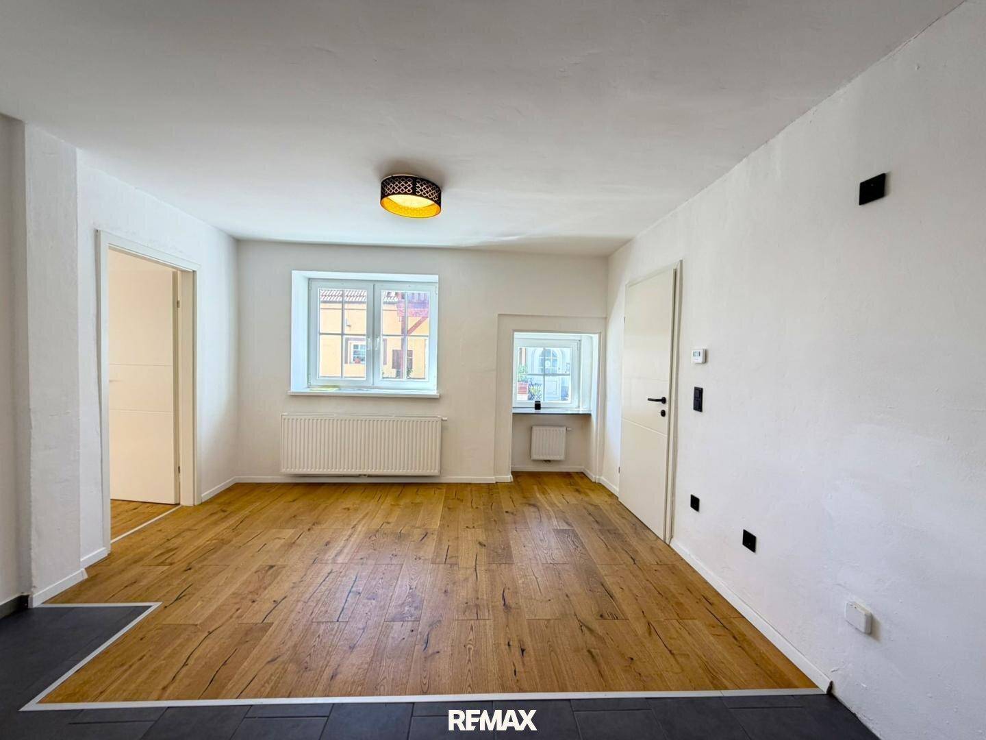 Heller Wohnraum mit Holzboden, weißen Wänden und zwei Fenstern mit Heizkörpern.