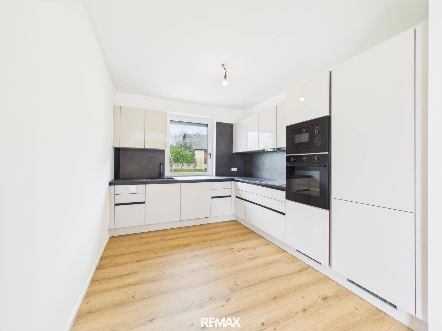 Moderne, leere Küche mit weißen Hochglanzschränken, dunkler Arbeitsplatte und Fenster.