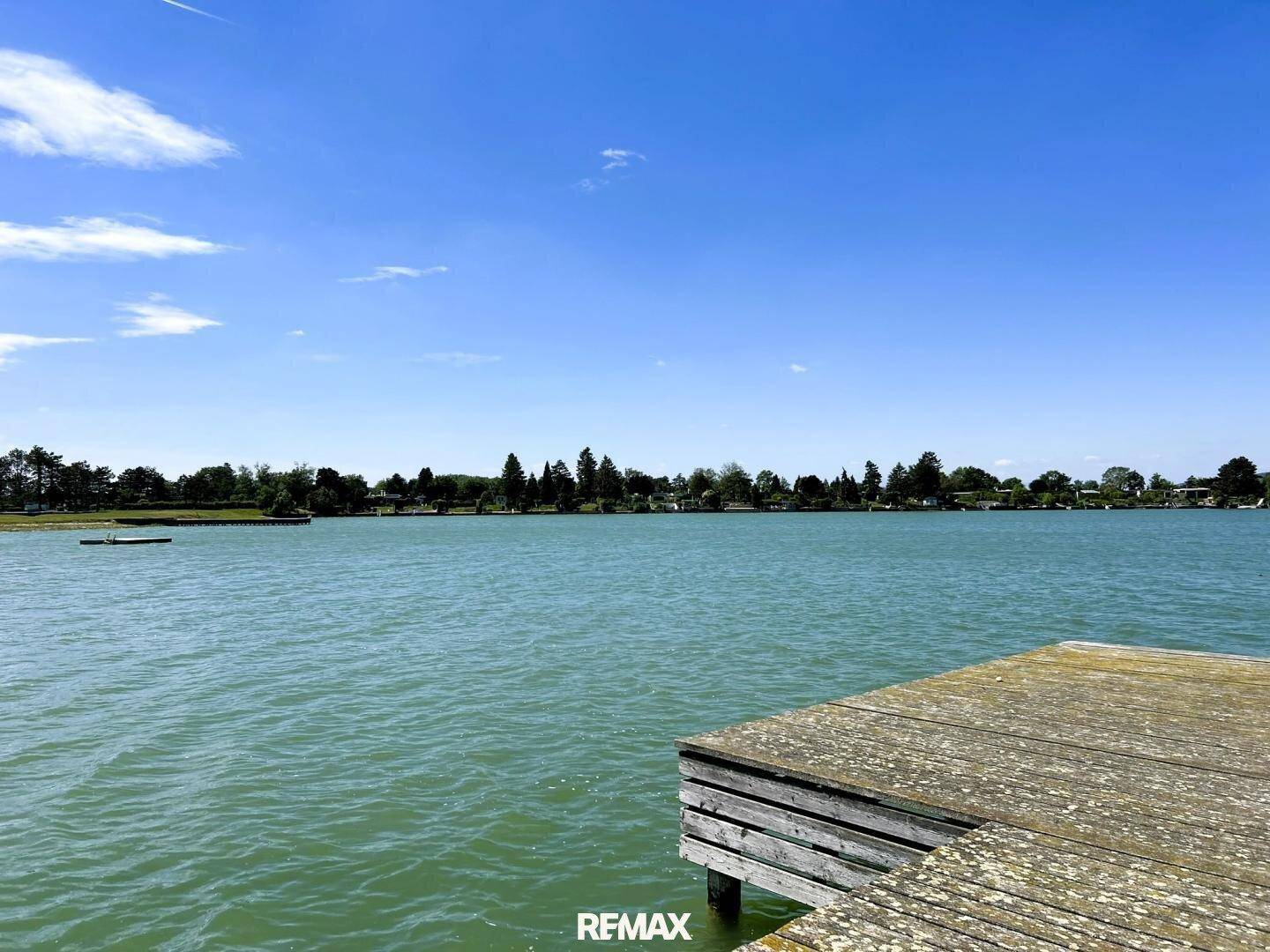 Weitläufiger Blick über den See von einem Holzsteg aus unter blauem Himmel.