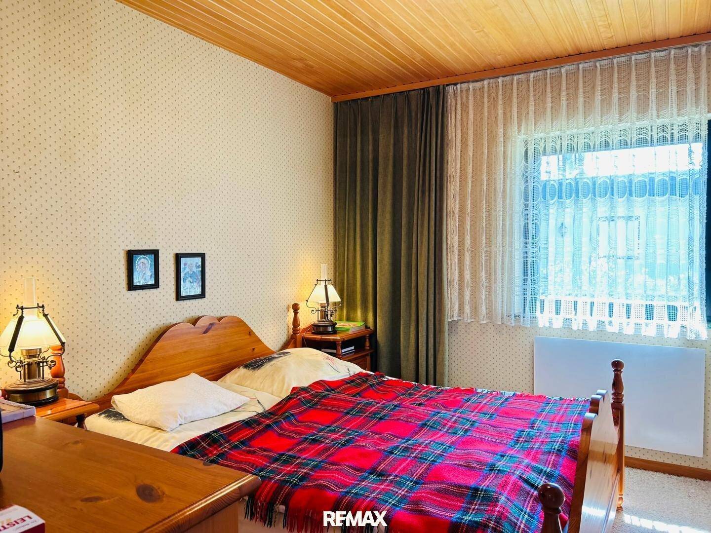 Schlafzimmer mit Holzbett, Holzdecke und gemusterter Tapete, hell durch das Fenster.