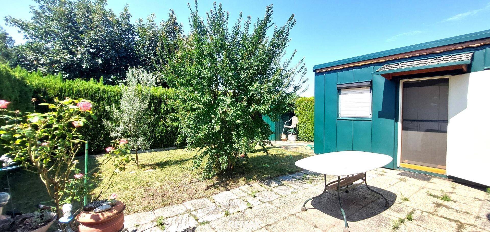 Sonniger Gartenbereich mit gepflasterter Fläche, einem weißen Gartentisch und blühenden Pflanzen.