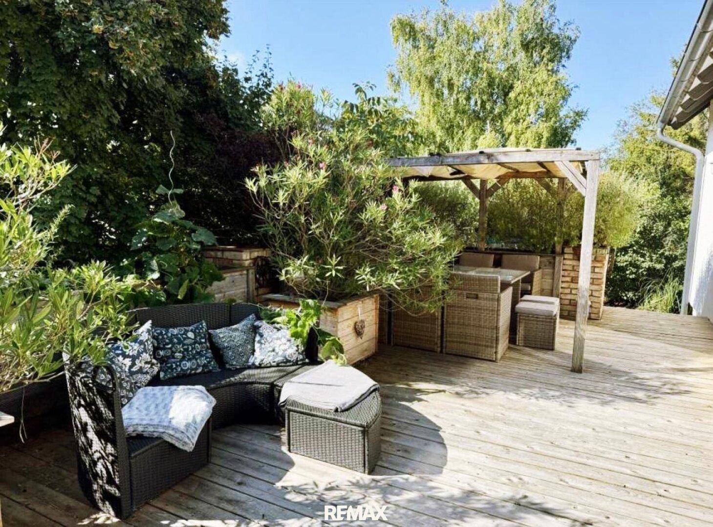 Geräumige Holzterrasse mit gemütlicher Lounge-Ecke und Essbereich unter einer Pergola im Grünen.