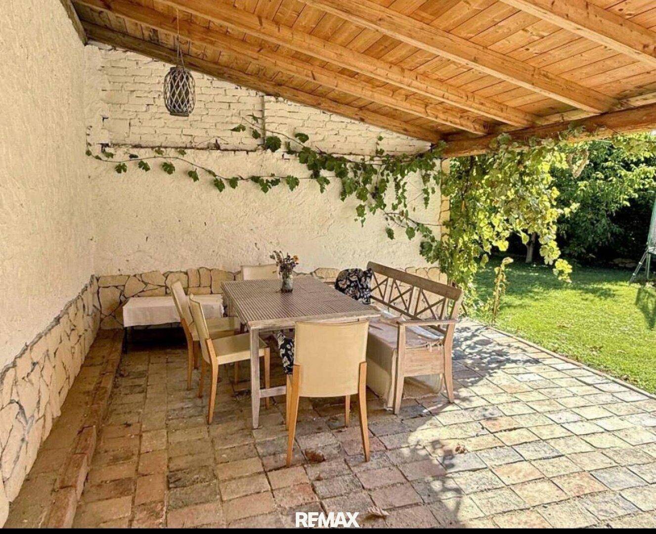 Überdachte Terrasse mit rustikalem Essbereich, Fliesenboden und Weinreben an der Wand.