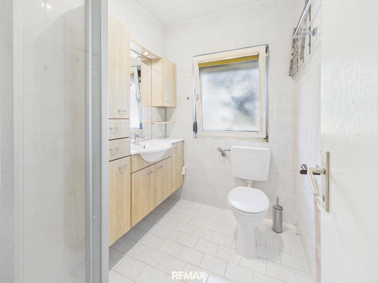 Helles Badezimmer mit Dusche, WC, Waschbecken und Holzschränken, gefliest in Weiß.