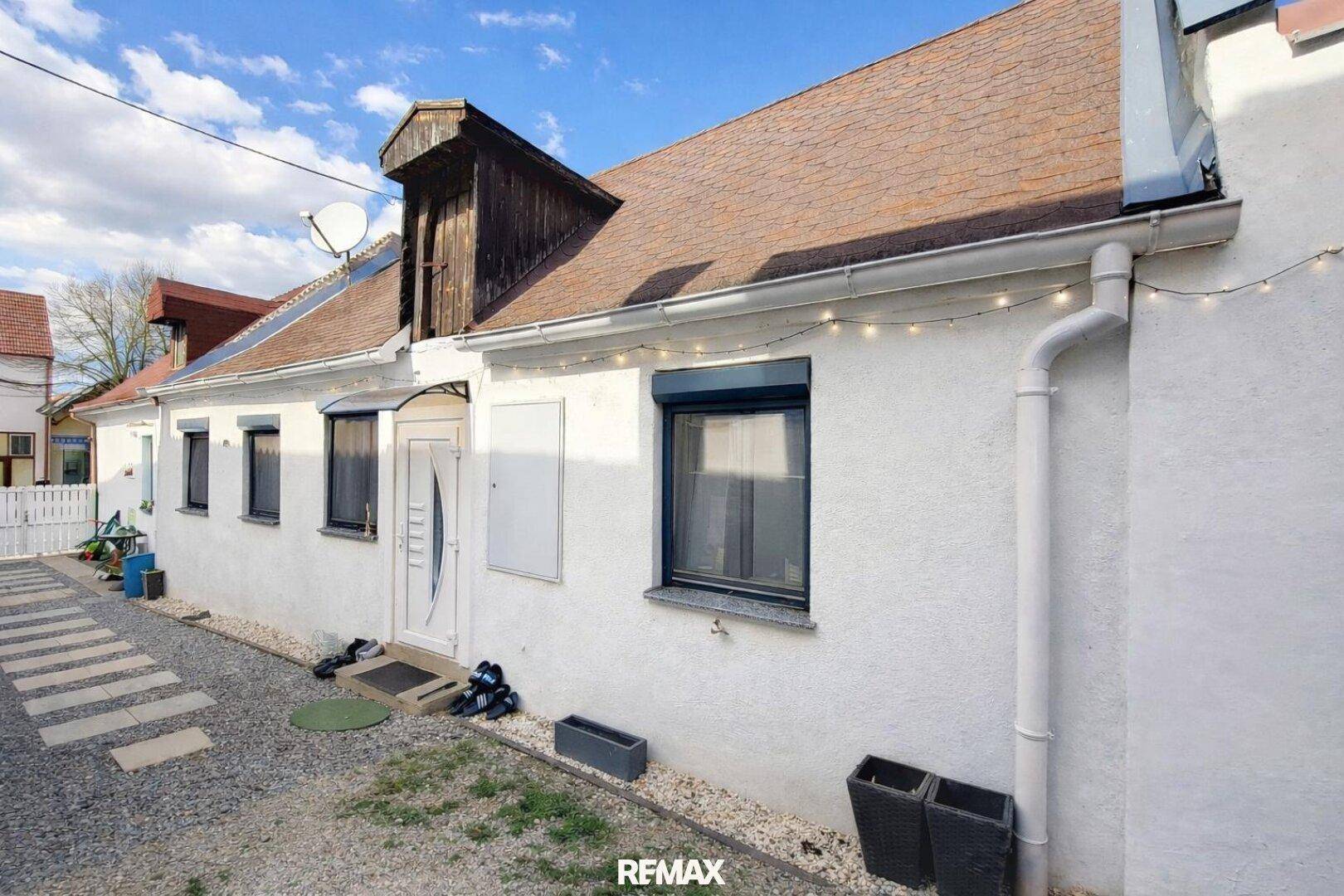 Weiße Hausfassade mit braunem Dach und einem kleinen Dachgauben, umgeben von einem Kiesweg.